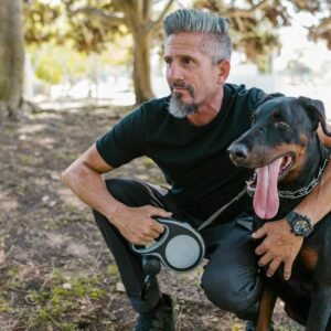Man crouching with a Doberman in a sunlit park, enjoying a stroll.