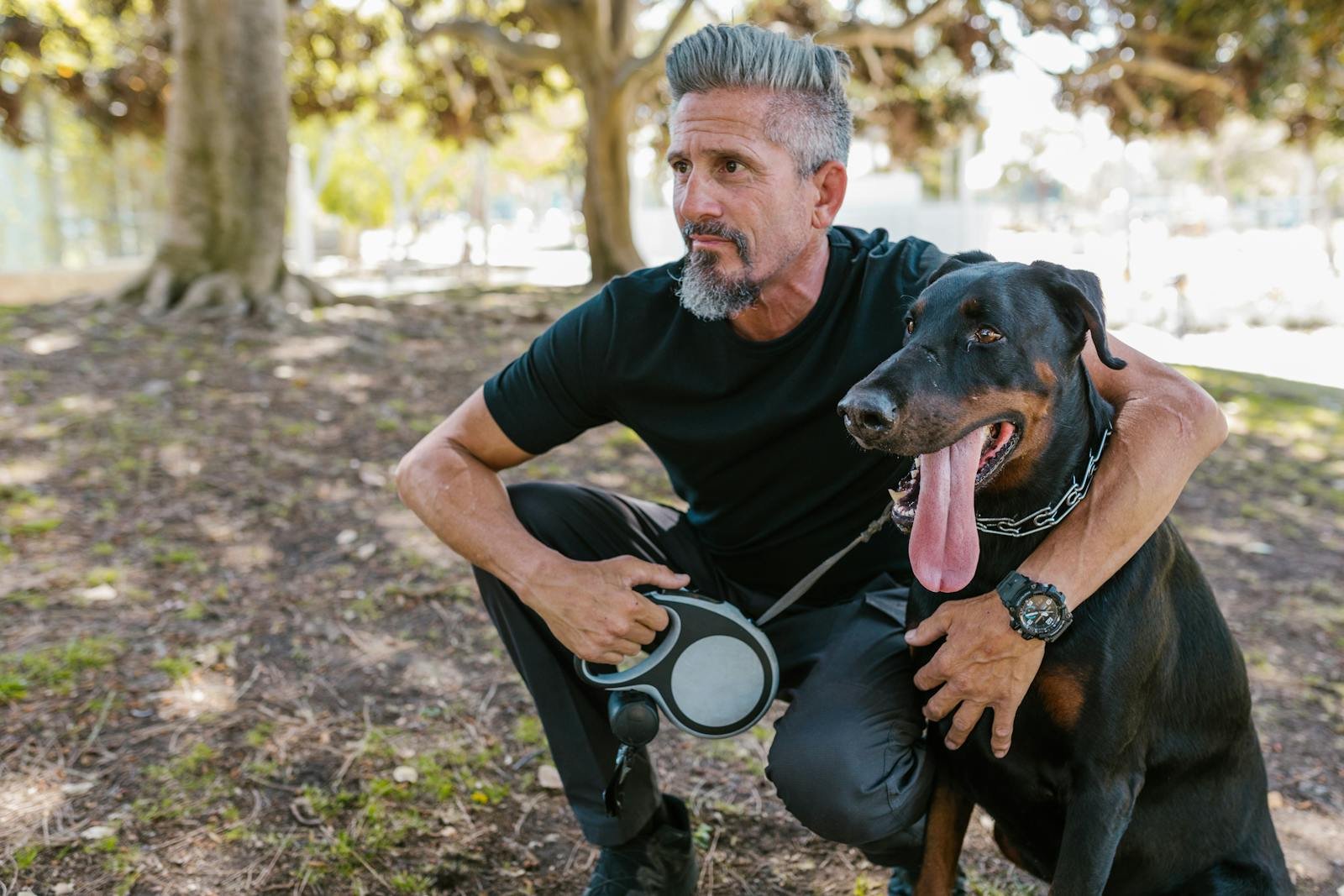 Man crouching with a Doberman in a sunlit park, enjoying a stroll.